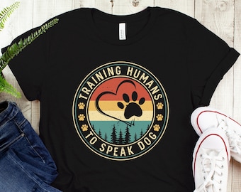 May include: Black t-shirt with a vintage-style graphic that says "Training Humans to Speak Dog". The graphic features a paw print inside a heart with a forest scene behind it.