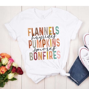 May include: White t-shirt with a graphic design featuring the words "Flannels", "Hayrides", "Pumpkins", "Smores", and "Bonfires" in colorful, textured blocks.