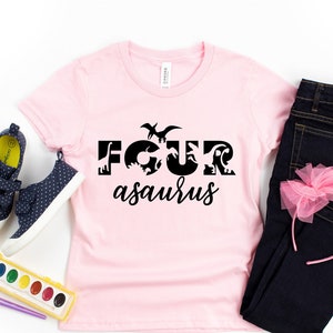 May include: Pink t-shirt with a black graphic that says "FOURasaurus" with dinosaur silhouettes forming the letters. The shirt is laid out on a white surface with a pair of blue sneakers, a set of watercolors, and a pair of blue jeans with a pink bow headband.