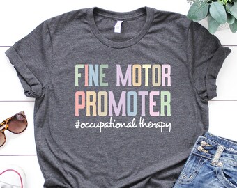 Pode incluir: Uma camiseta cinza escura com o texto "FINE MOTOR PROMOTER #occupational therapy" em cores do arco-íris. A camiseta está colocada sobre uma superfície de madeira branca com óculos de sol, um par de tênis brancos e uma calça jeans azul.