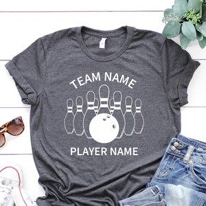 May include: A gray t-shirt with a white bowling pin graphic and the text "TEAM NAME" and "PLAYER NAME".