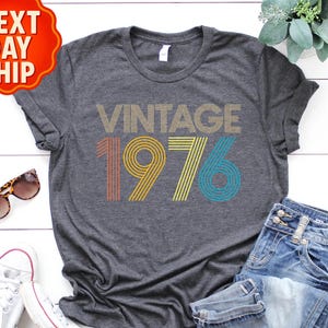 May include: Dark gray t-shirt with the text "VINTAGE 1976" in a retro design. The shirt is paired with blue jeans, white sneakers, and sunglasses. A red and orange "NEXT DAY SHIP" sticker is in the top left corner.