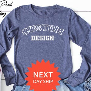 May include: A blue long-sleeve shirt with the words "CUSTOM DESIGN" in white lettering. The shirt is paired with blue jeans and white sneakers. The image also includes the text "NEXT DAY SHIP".