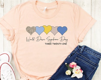 May include: A peach-colored t-shirt with a row of five colorful hearts and the text "World Down Syndrome Day THREE TWENTY-ONE". The shirt is laid out on a wooden surface with a pair of white sneakers and blue jeans.