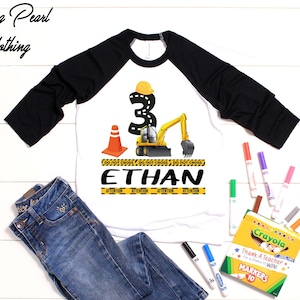 May include: White and black raglan shirt with a construction-themed design. Features a yellow excavator, the number 3, and the name "Ethan". Includes blue jeans and a box of Crayola markers.