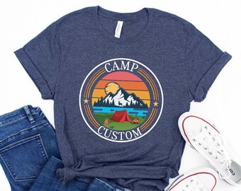 May include: A navy blue t-shirt with a vintage-style graphic of a mountain scene with a red tent, a car, and a bear. The graphic is surrounded by a circle with the text "CAMP CUSTOM" in white.