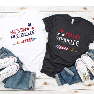 May include: Two t-shirts with a Fourth of July theme. The white t-shirt says "She's my Firecracker" with a red, white and blue firework graphic. The dark grey t-shirt says "I'm his Sparkler" with a red, white and blue sparkler graphic.