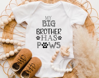 May include: White baby onesie with black text that reads "My Big Brother Has Paws" with paw prints. The onesie is on a woven wicker surface with a wooden bead necklace and brown leather baby shoes.