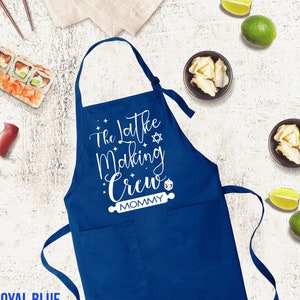 May include: Royal blue apron with the text "The Latke Making Crew Mommy" in white script. The apron has a large front pocket and adjustable neck strap. Sushi, limes, and small bowls of food are arranged around the apron.