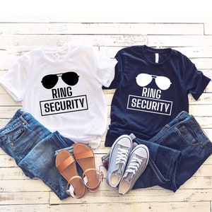 May include: Two t-shirts with the text "Ring Security" and a pair of sunglasses graphic. The shirts are white and navy blue. The shirts are laid out on a white wood background with a pair of blue jeans, a pair of brown sandals, and a pair of gray sneakers.