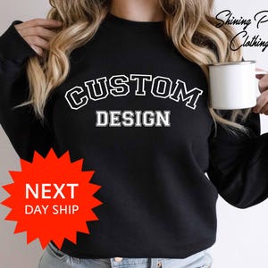 May include: Black sweatshirt with the words "CUSTOM DESIGN" in white, collegiate-style lettering. The sweatshirt is plain, with a crew neck. A white mug is held in the right hand. The words "Shining Pearl Clothing" are in the upper right corner.
