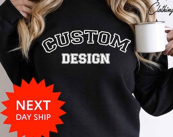 May include: Black sweatshirt with the words "CUSTOM DESIGN" in white, collegiate-style lettering. The sweatshirt is plain, with a crew neck. A white mug is held in the right hand. The words "Shining Pearl Clothing" are in the upper right corner.