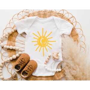 Pode incluir: Um macac&atilde;o branco para beb&ecirc; com um gr&aacute;fico de sol amarelo. O macac&atilde;o est&aacute; sobre uma superf&iacute;cie de vime tecida com um colar de contas de madeira e um mordedor de madeira.
