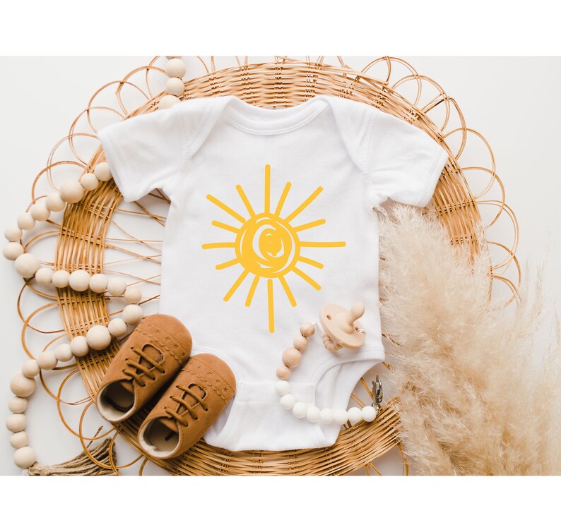 Pode incluir: Um macac&atilde;o branco para beb&ecirc; com um gr&aacute;fico de sol amarelo. O macac&atilde;o est&aacute; sobre uma superf&iacute;cie de vime tecida com um colar de contas de madeira e um mordedor de madeira.
