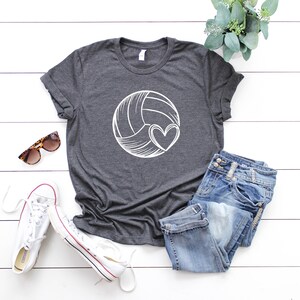 May include: A dark gray t-shirt with a white volleyball and heart design. The shirt is laid out on a white wooden surface with a pair of sunglasses, a pair of white sneakers, and a pair of blue jeans.