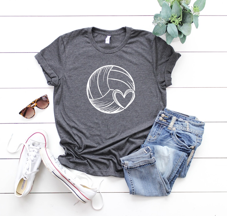 May include: A dark gray t-shirt with a white volleyball and heart design. The shirt is laid out on a white wooden surface with a pair of sunglasses, a pair of white sneakers, and a pair of blue jeans.