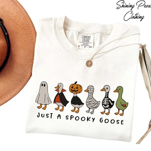 A white t-shirt with a graphic design of five cartoon ducks dressed in Halloween costumes, including a ghost, skeleton, and mummy, along with the text "Just a Spooky Goose".