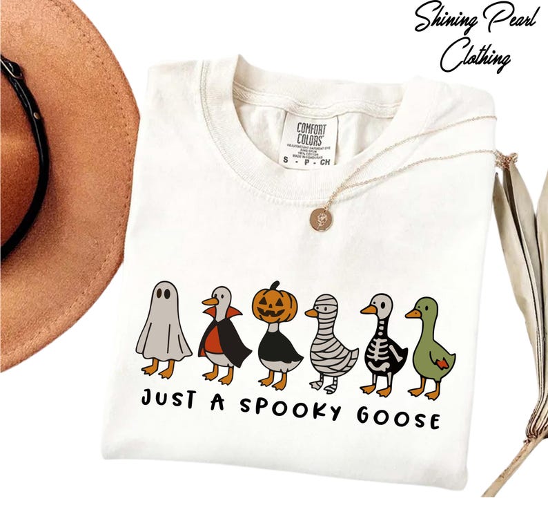 A white t-shirt with a graphic design of five cartoon ducks dressed in Halloween costumes, including a ghost, skeleton, and mummy, along with the text "Just a Spooky Goose".