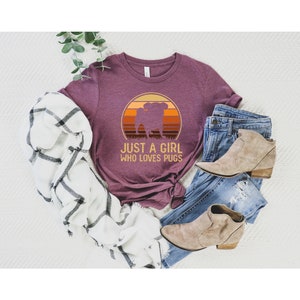 Peut inclure: Un t-shirt marron avec un graphique vintage d'un carlin devant un coucher de soleil. Le texte "Just a girl who loves pugs" est imprim&eacute; sous le graphique.