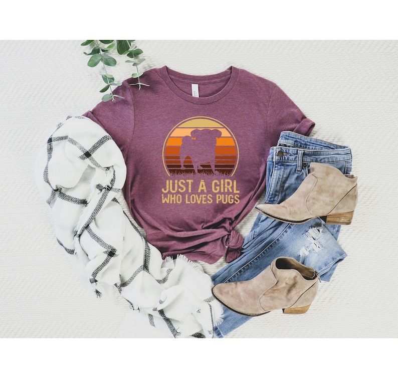 Peut inclure: Un t-shirt marron avec un graphique vintage d'un carlin devant un coucher de soleil. Le texte "Just a girl who loves pugs" est imprim&eacute; sous le graphique.