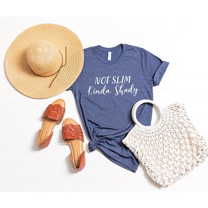 Puede incluir: Una camiseta azul marino con el texto "Not Slim Kinda Shady" en letras blancas. La camiseta est&aacute; colocada sobre una superficie de madera blanca con un sombrero de paja, sandalias marrones y un bolso de crochet blanco.