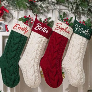 May include: Four cable-knit Christmas stockings hang against a white door. Each stocking is a different color: green, white, red, and white. The names "Emily", "Bella", "Sophia", and "Martin" are embroidered on the cuffs.
