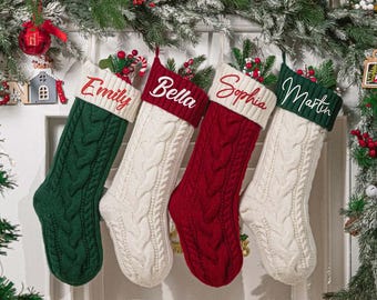 May include: Four cable-knit Christmas stockings hang against a white door. Each stocking is a different color: green, white, red, and white. The names "Emily", "Bella", "Sophia", and "Martin" are embroidered on the cuffs.