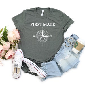 May include: A green t-shirt with the text "FIRST MATE" and a compass graphic. The shirt is laid out on a white background with a pair of blue jeans, a pair of white sneakers, and a bouquet of pink flowers.