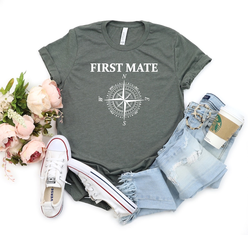 May include: A green t-shirt with the text "FIRST MATE" and a compass graphic. The shirt is laid out on a white background with a pair of blue jeans, a pair of white sneakers, and a bouquet of pink flowers.