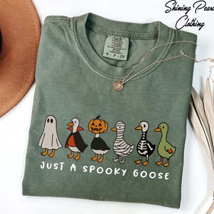A green t-shirt with a Halloween-themed graphic of a skeleton, pumpkin, ghost, and a goose, along with the text "Just a spooky goose" printed on it.