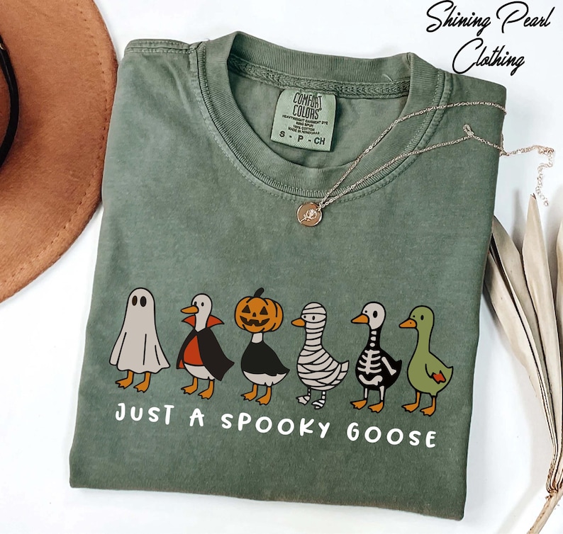 A green t-shirt with a Halloween-themed graphic of a skeleton, pumpkin, ghost, and a goose, along with the text "Just a spooky goose" printed on it.