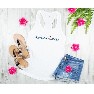 May include: White tank top with a blue cursive word "america" and a red star. The tank top is lying on a white wooden surface with pink flowers and a pair of sandals.