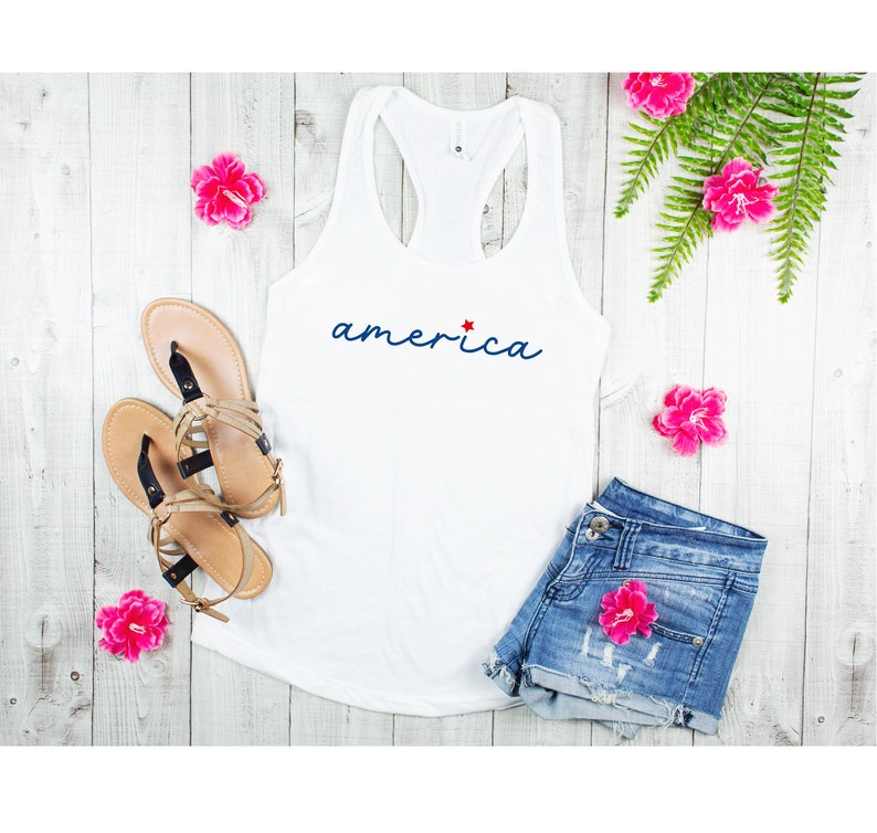 May include: White tank top with a blue cursive word "america" and a red star. The tank top is lying on a white wooden surface with pink flowers and a pair of sandals.