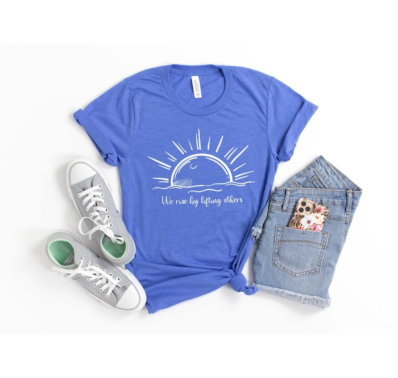 May include: A royal blue t-shirt with a white graphic of a sun rising over the ocean. The text "We rise by lifting others" is printed below the graphic.
