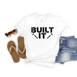 Puede incluir: Camiseta blanca con texto negro que dice "BUILT IT" con dos martillos negros a cada lado del "IT".