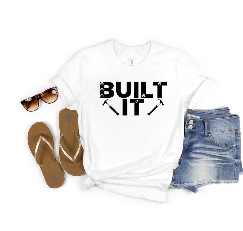 Puede incluir: Camiseta blanca con texto negro que dice "BUILT IT" con dos martillos negros a cada lado del "IT".