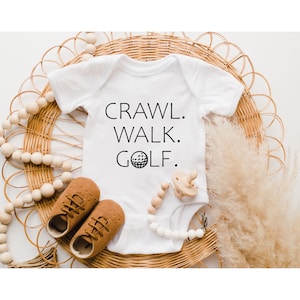 May include: White baby onesie with black text that reads "Crawl. Walk. Golf." and a golf ball graphic.