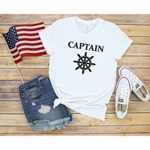 May include: White t-shirt with the word "CAPTAIN" and a black ship's wheel graphic. The shirt is laid out on a wooden surface with a pair of white sneakers, a pair of denim shorts, and an American flag.