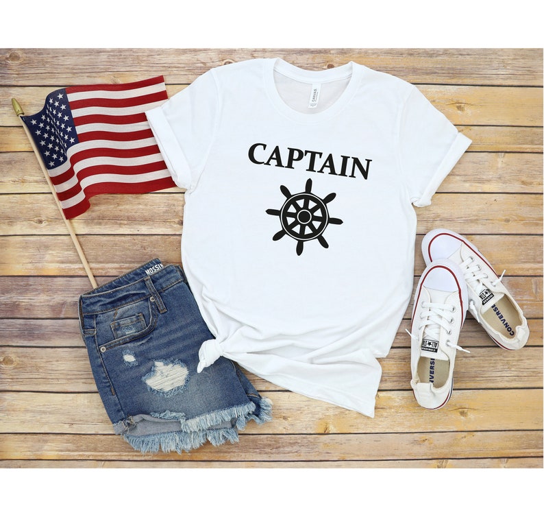 May include: White t-shirt with the word "CAPTAIN" and a black ship's wheel graphic. The shirt is laid out on a wooden surface with a pair of white sneakers, a pair of denim shorts, and an American flag.