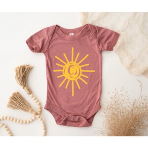 Pode incluir: Um macac&atilde;o de beb&ecirc; malva com um gr&aacute;fico de sol amarelo. O sol &eacute; desenhado em um estilo simples e infantil.