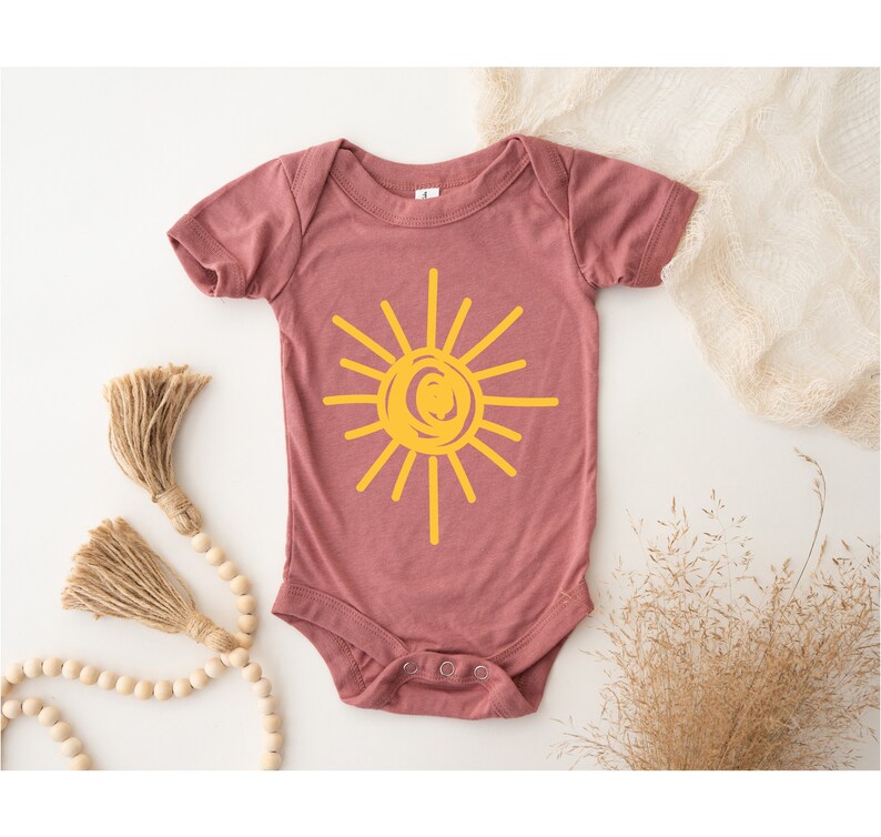 Pode incluir: Um macac&atilde;o de beb&ecirc; malva com um gr&aacute;fico de sol amarelo. O sol &eacute; desenhado em um estilo simples e infantil.