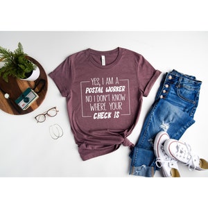 May include: A maroon t-shirt with white text that reads "YES, I AM A POSTAL WORKER NO I DON'T KNOW WHERE YOUR CHECK IS". The shirt is laid out on a white surface with a pair of jeans, a pair of white sneakers, and a potted plant.