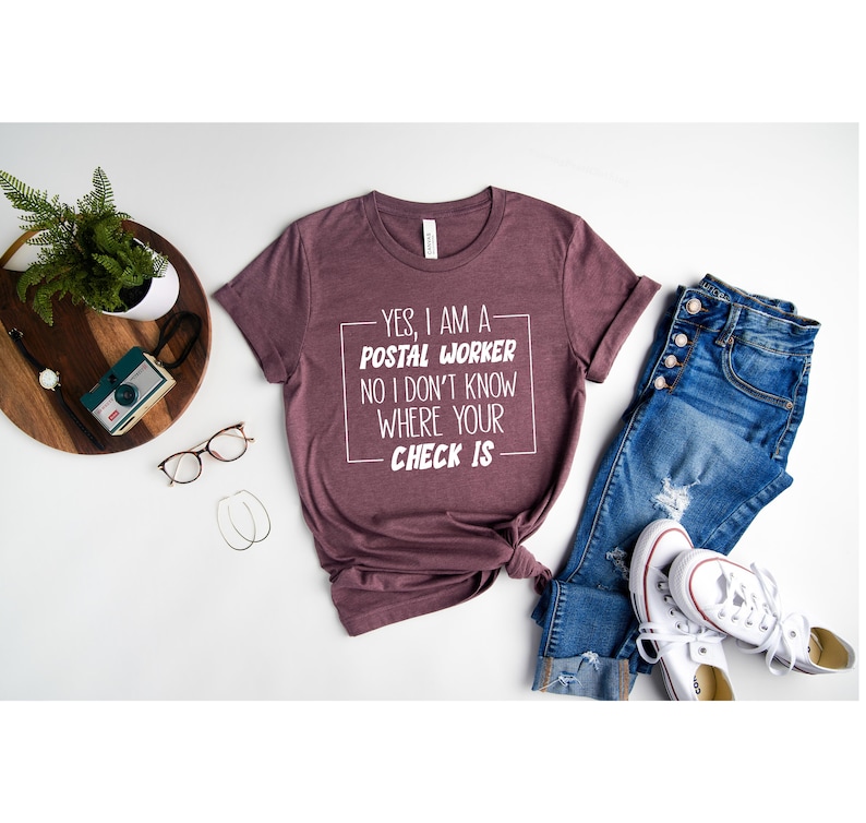May include: A maroon t-shirt with white text that reads "YES, I AM A POSTAL WORKER NO I DON'T KNOW WHERE YOUR CHECK IS". The shirt is laid out on a white surface with a pair of jeans, a pair of white sneakers, and a potted plant.