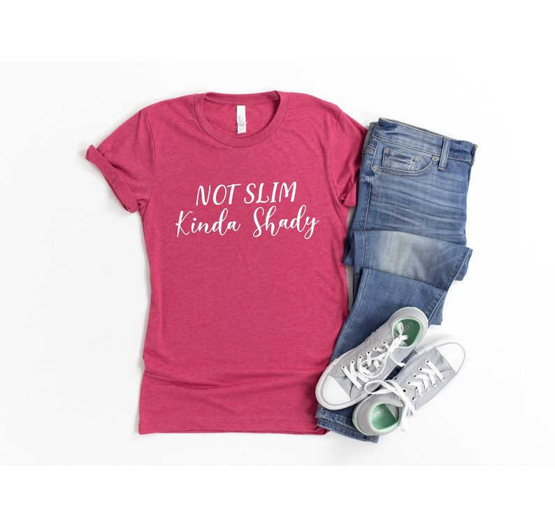 Puede incluir: Una camiseta roja con el texto "NOT SLIM Kinda Shady" en letras blancas. La camiseta se combina con unos vaqueros azules y zapatillas grises.