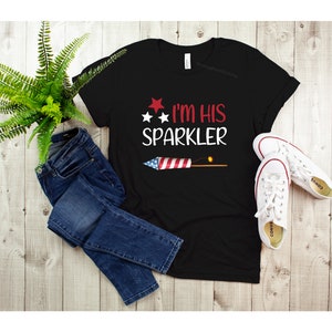 May include: Black t-shirt with a red, white and blue firework graphic and the text "I'm His Sparkler".