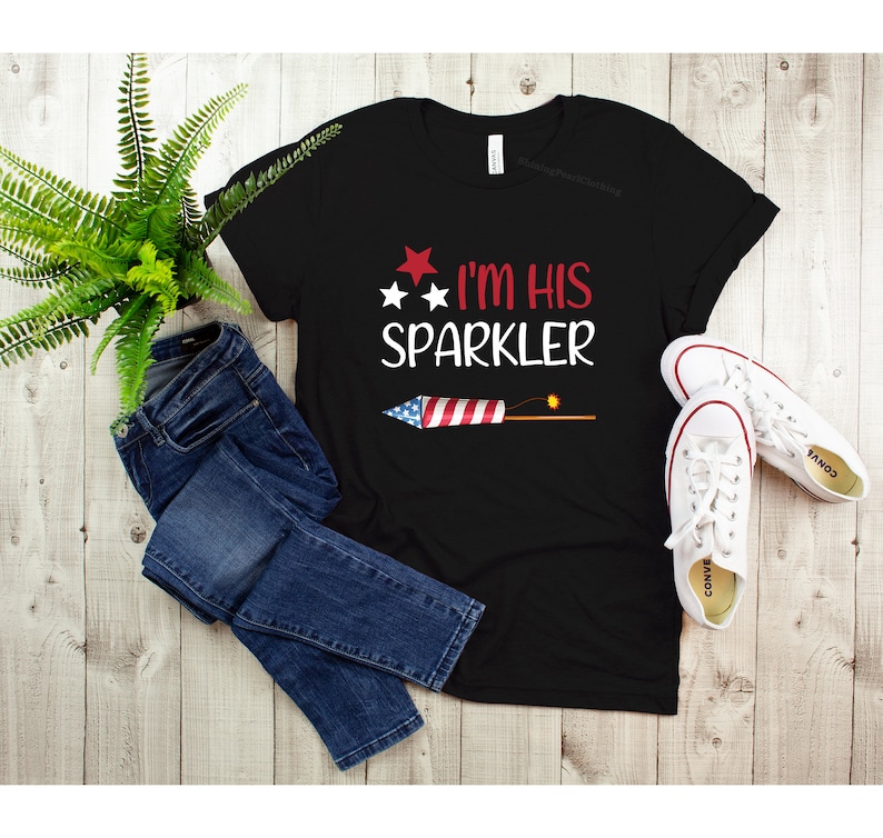 May include: Black t-shirt with a red, white and blue firework graphic and the text "I'm His Sparkler".