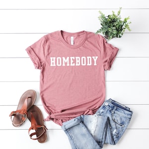 May include: A dusty rose t-shirt with the word "HOMEBODY" printed in white on the front. The shirt is laid out on a white wooden surface with a pair of brown sandals and blue jeans.