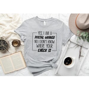 May include: A gray t-shirt with black text that reads "YES, I AM A POSTAL WORKER NO I DON'T KNOW WHERE YOUR CHECK IS".
