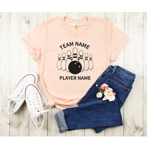 May include: Peach colored t-shirt with black text that reads "TEAM NAME" and "PLAYER NAME" with a graphic of bowling pins and a bowling ball.