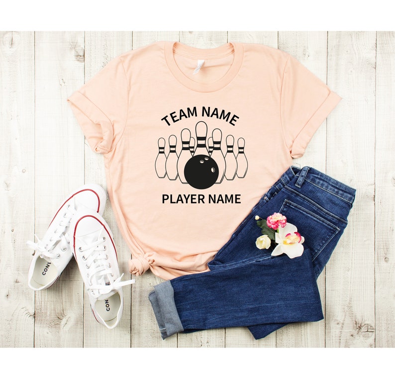 May include: Peach colored t-shirt with black text that reads "TEAM NAME" and "PLAYER NAME" with a graphic of bowling pins and a bowling ball.